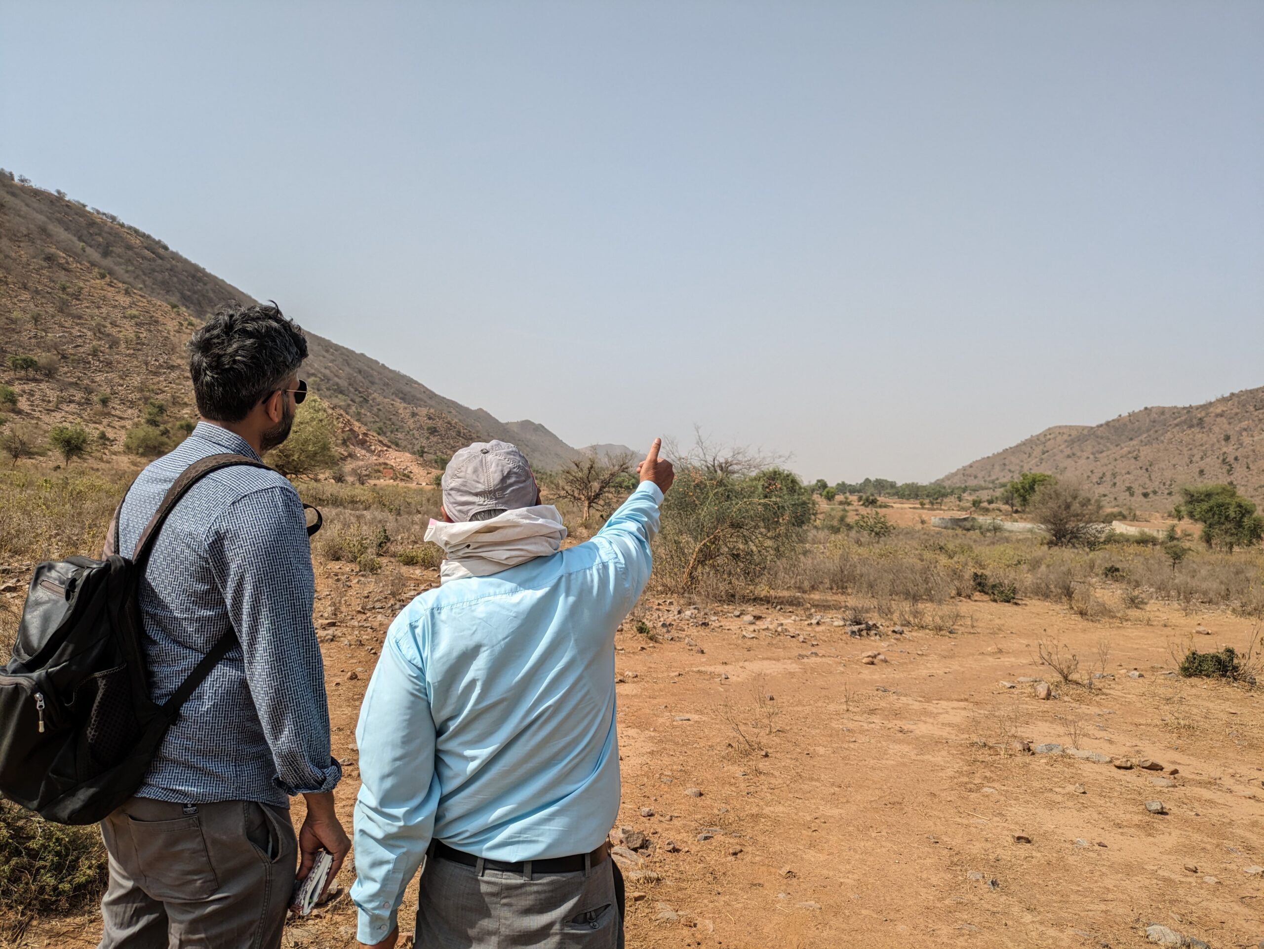 Two men standing on the left side of the image. They face away from the camera. A man wearing a cap and a towel around his neck points at a distance, while a man wearing a bag pack and sunglasses looks on. The background is of an arid landscape.