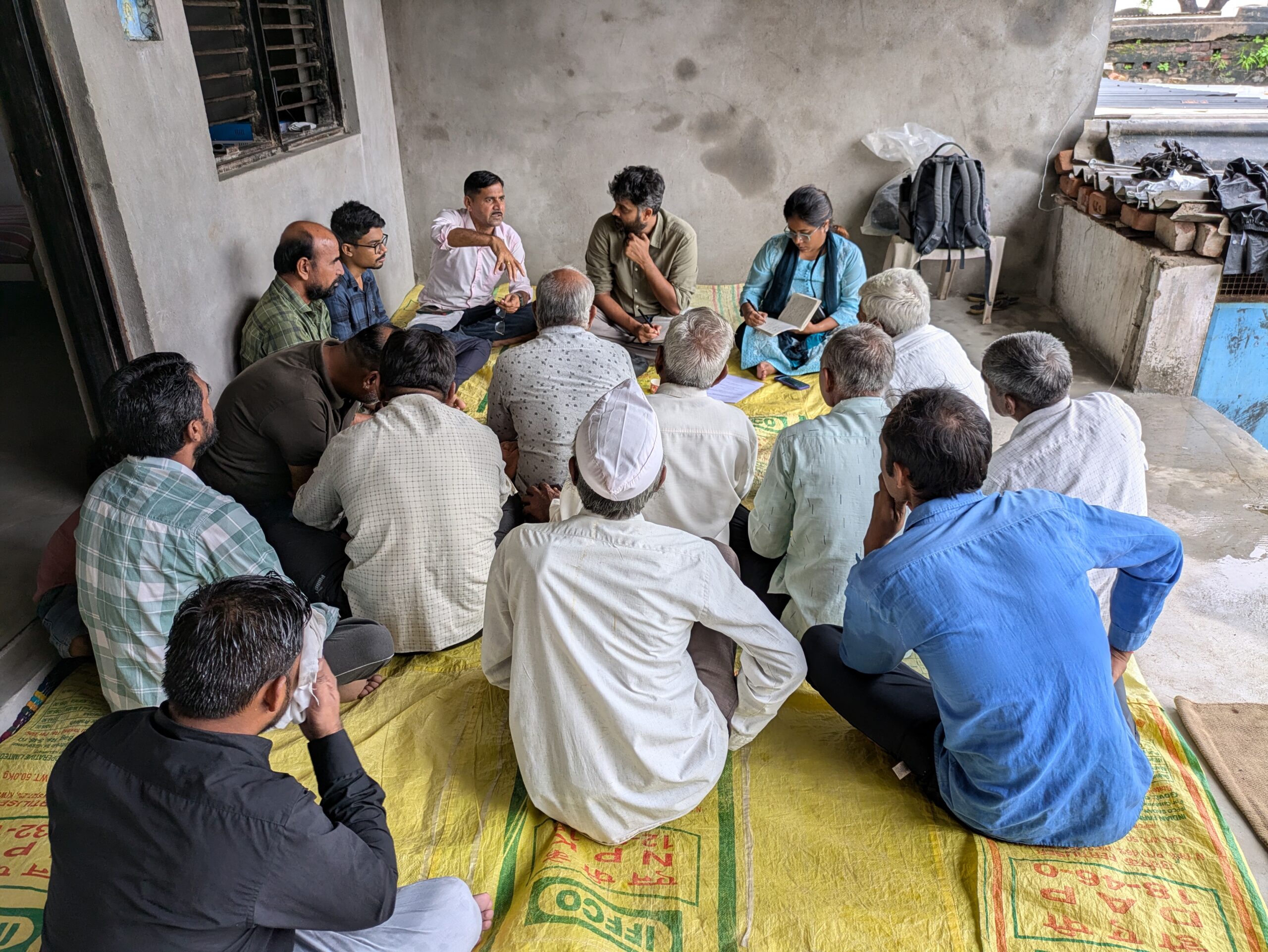 A group of about 15 people sitting on a yellow plastic mat, speaking to each other. Some are making notes. The space is closed on three sides, with bare cemented walls.