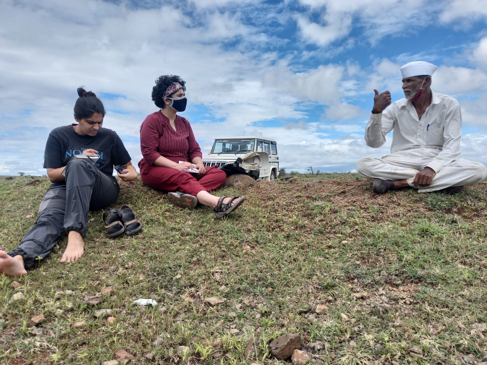 Image of two women on the left carrying notebooks and writing in them, speaking to a man on the right. They are sitting on dry, open land. The sky is clear and a jeep is parked behind them.